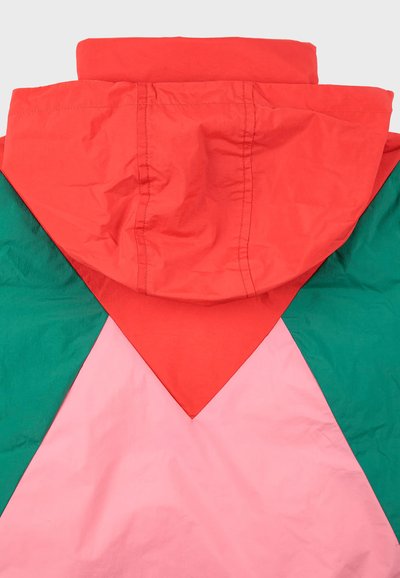 Veste à blocs de couleurs avec une capuche rouge, du tissu vert et rose dans un motif géométrique, faite d'un matériau léger et légèrement froissé.