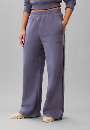 Persoon die losse lavendelkleurige joggingbroek draagt met elastische tailleband en twee zijzakken, gecombineerd met beige schoenen op een grijze achtergrond.