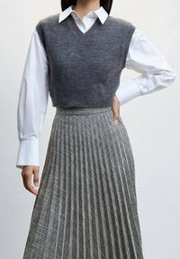 Gilet grigio lavorato a maglia sopra una camicia bianca a maniche lunghe, abbinato a una gonna pieghettata grigia e nera a motivi. Design ordinato e strutturato.
