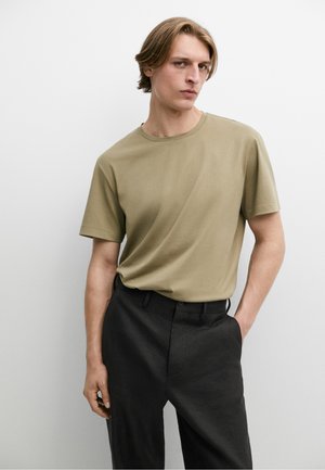 Giovane uomo con capelli castano chiaro che indossa una maglietta beige a maniche corte infilata in pantaloni neri, in piedi davanti a uno sfondo bianco uniforme.