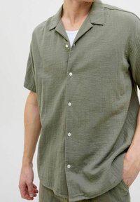 Korte mouwen olijfgroene shirt van gestructureerde stof, met een kraag en witte knopen aan de voorkant.