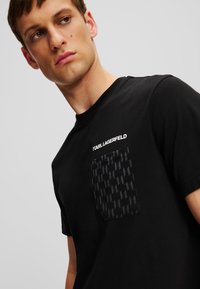 Melnas kokvilnas t-krekls ar apaļu kaklu, ar teksturētu kabatu, kurā ir tumša raksts, un "KARL LAGERFELD" logo, kas izsprints virs kabatas.