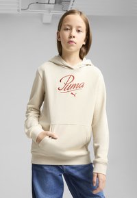 Felpa beige con logo rosso "Puma", caratterizzata da una tasca frontale e cappuccio con cordino. Indossata con jeans in denim blu. Tessuto liscio e casual.