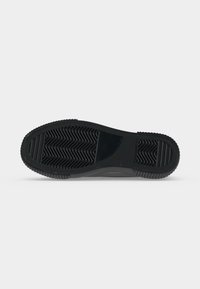 Schwarze Sneaker-Sohle mit strukturiertem Grip und Zickzack- sowie Streifenmustern auf einem schlichten hellen Hintergrund.