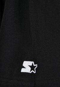 Melna kokvilnas auduma ar gludu tekstūru, kas izmanto baltu izšūtu logo, kurā ir "S" burts un zvaigzne pie apakšmalas.