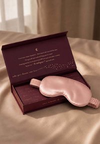 Masque pour les yeux en soie rose reposant sur une boîte ouverte violette foncée avec un texte doré à l'intérieur, posé sur une surface en tissu beige.