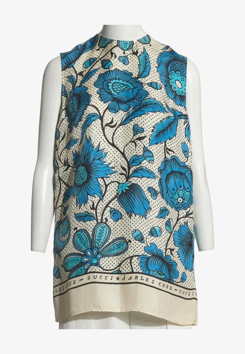 Haut beige sans manches avec grands motifs floraux bleus et branches noires, présenté sur un torse de mannequin blanc.