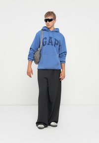 Joven con una sudadera azul de GAP, pantalones negros holgados, bolso cruzado gris, zapatillas gris y gafas de sol negras de pie frente a un fondo blanco.