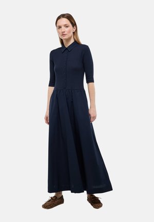 Femme portant une robe maxi bleu marine boutonnée avec des manches trois-quarts et des chaussures imprimé léopard, debout devant un fond clair uni.