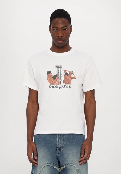 Homme portant un T-shirt blanc avec un motif d'hommes torse nu et le texte "Sounds gay, I'm in", associé à un jean bleu aux genoux déchirés.