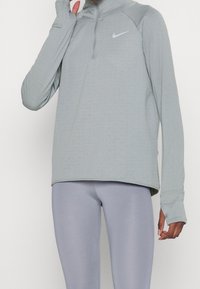 Ljusgrå, långärmad sporttröja med halvlång dragkedja, ribbad textur och tumhål. Matcher med släta, ljusgrå leggings.