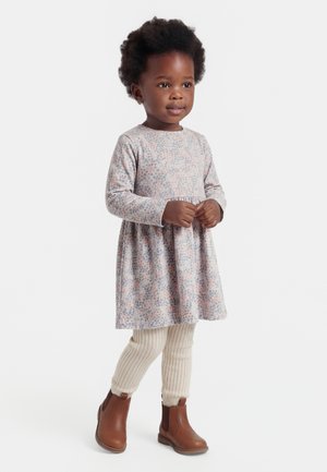 Young child standing, wearing a long-sleeve floral dress, cream ribbed leggings, and brown ankle boots against a white background.