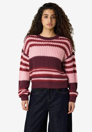 Femme aux longs cheveux bouclés portant un pull en tricot rayé rose, bordeaux et rouge et un jean bleu foncé, debout devant un fond uni.