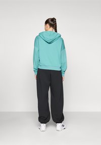 Turkis hettegensere med hette og senket skuldre, kombinert med løse svarte joggebukser og hvite treningssko. Glatt stofftekstur.
