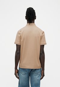 Άνδρας με σκουρόχρωμο δέρμα και πλεξούδες, εικονιζόμενος από πίσω, φορώντας μπεζ μπλουζάκι πόλο με κοντά μανίκια και μπλε τζιν, μπροστά σε απλό λευκό φόντο.