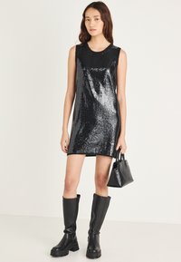 Μαύρο φόρεμα με παγιέτες σε ίσια γραμμή, στρογγυλή λαιμόκοψη, αμάνικο, σε συνδυασμό με μαύρες μπότες μέχρι το γόνατο και μικρή μαύρη τσάντα.