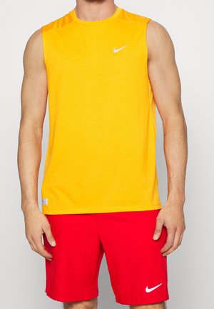Homme portant un débardeur de sport jaune et un short de sport rouge avec des logos blancs Nike, debout devant un fond uni.