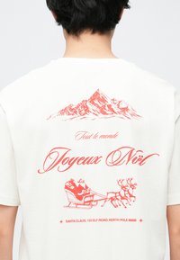 Hvid bomuld T-shirt med rødt grafisk design af bjerge, "Joyeux Noël" og en slæde med Julemanden og rensdyr på bagsiden.