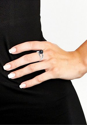 Bague en argent ornée d'une pierre noire ronde, montée sur un anneau au design complexe, portée sur une main aux ongles clairs devant un fond sombre.
