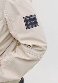 Veste softshell beige avec une finition texturée, dotée d'une étiquette carrée noire sur la manche avec des détails de texte blanc et un logo.