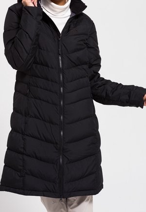 Persoon draagt een lange zwarte doorgestikte donzen jas met een ritssluiting aan de voorkant over een witte coltrui.