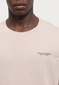 T-shirt di cotone beige chiaro con scollo rotondo. Presenta un piccolo logo grafico scuro nella parte sinistra del petto. Texture liscia.