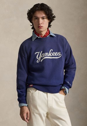 Sweat-shirt bleu marine avec broderie "Yankees" en blanc, associé à une chemise en denim bleu clair. Pantalon blanc et bandana rouge complètent la tenue.