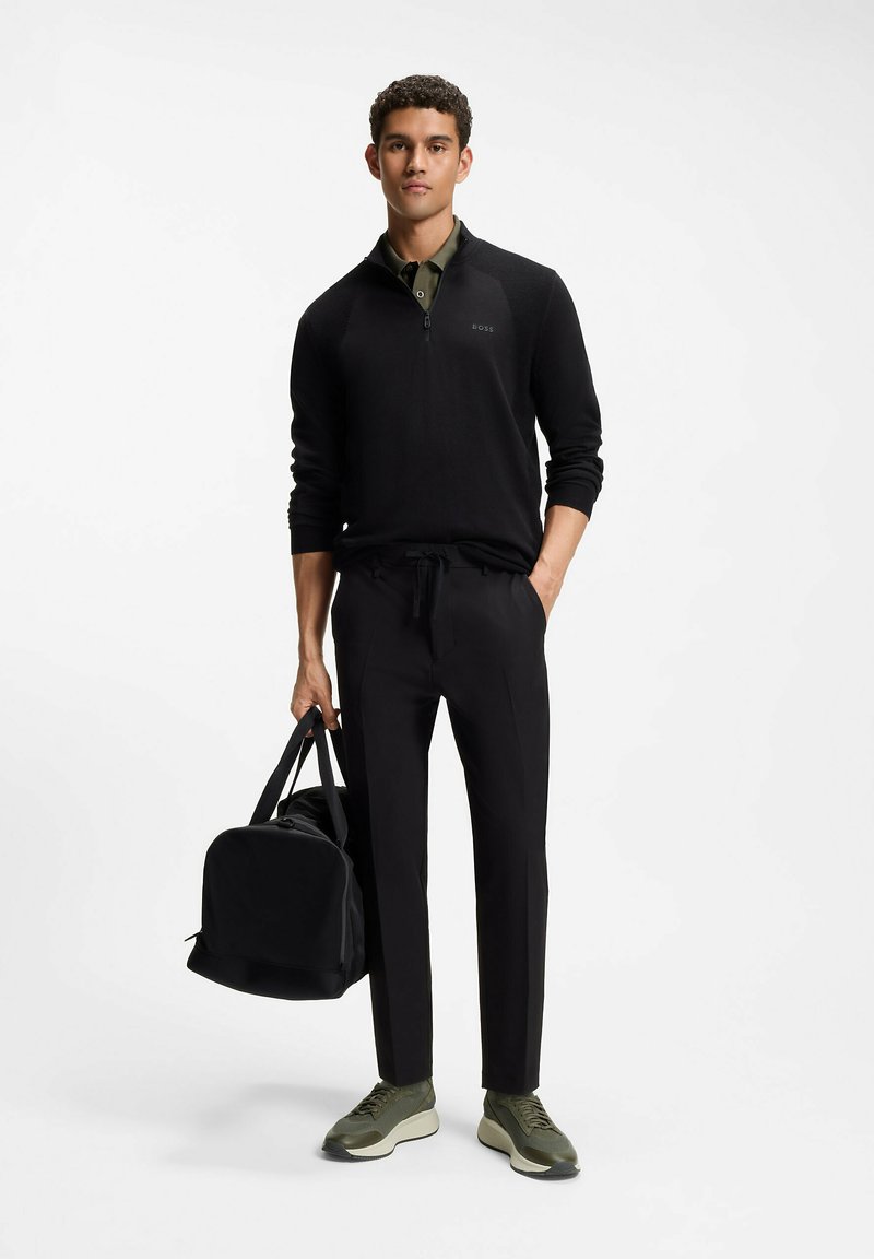 Jeune homme en pull zippé noir, pantalon noir et baskets vertes tenant un sac de sport noir avec une main dans la poche.