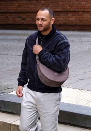 Hombre con chaqueta negra y pantalones claros caminando al aire libre, llevando un bolso marrón cruzado en el hombro.