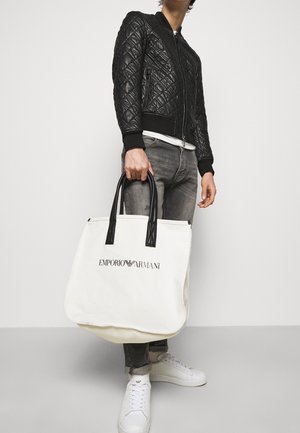 Personne portant une veste matelassée noire, un jean gris, des baskets blanches, tenant un grand sac fourre-tout blanc Emporio Armani avec des poignées noires.