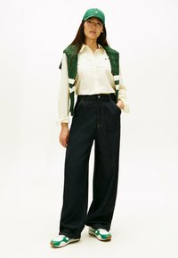 Groene gewatteerde bodywarmer over een wit gestreept shirt, gecombineerd met high-waisted donker denimbroek en groen-witte sneakers. Groene pet op het hoofd.