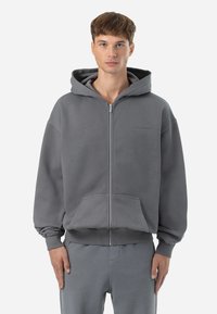 Felpa grigia con zip realizzata in tessuto morbido, dotata di una tasca frontale, cappuccio con lacci e polsini e orlo a coste. Dettaglio del logo minimalista sul petto.