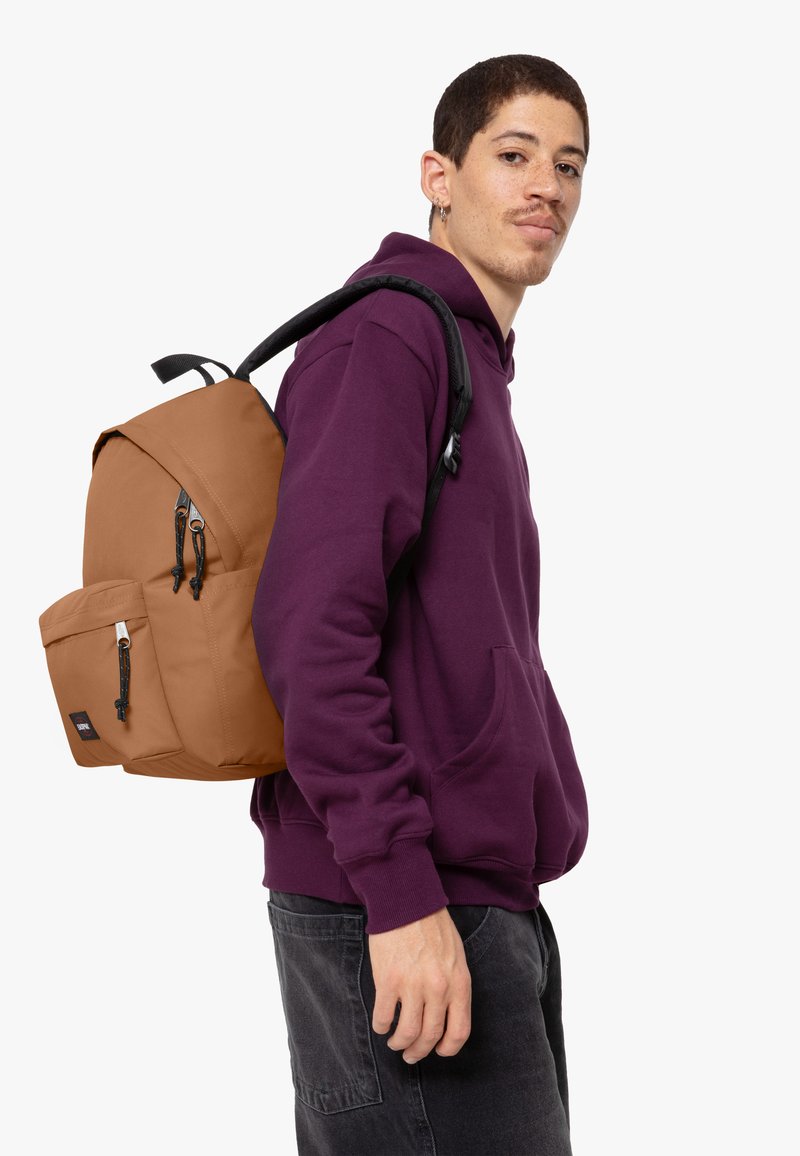 Zaino marrone con un grande scomparto con zip, tasca anteriore e spallacci neri regolabili. Felpa viola, pantaloni grigi e capelli corti.