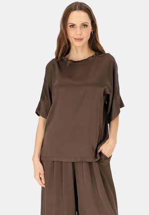 Kobieta ubrana w luźną ciemnobrązową bluzkę z krótkim rękawem i pasujące szerokie spodnie, stojąca z jedną ręką w kieszeni na jednolitym tle.