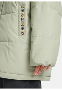 Helle grüne gepolsterte Jacke mit geometrischem Muster an der Taschenöffnung und einer Hand mit sichtbarem Tattoo nahe dem Saum vor weißem Hintergrund.