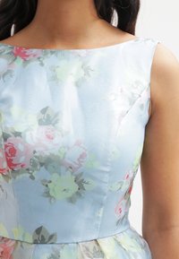 Vaaleansininen kukkamekko, jossa on sileä ja kiiltävä pinta. Siinä on pastellivärisiä ruusuja ja vihreitä lehtiä vesivärityylillä, hihaton malli.