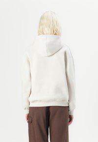 A person with shoulder-length, wavy blonde hair wears a light beige hoodie and brown cargo pants, facing away against a plain white background.