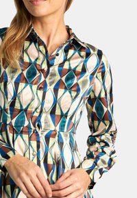 Vestido tipo camisa con un patrón geométrico en azul, verde, marrón y crema, con una cintura ajustada y mangas largas. Textura suave similar a la seda.