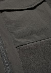 Mørkegrå fleecejakke-sektion med en lynlåslomme, tekstureret stof og præget logo-detalje, samt syninger langs kanterne.