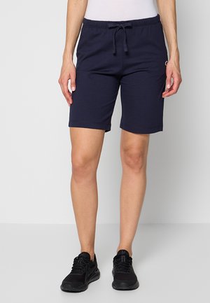 Personne portant un short bleu marine à cordon, un haut blanc et des chaussures de sport noires, debout devant un fond clair uni.