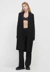 Schwarzer langer Cardigan mit offenem Vorderteil, aus weichem Material gefertigt. Verfügt über weite Ärmel und fällt locker über schwarze, figurbetonte Hosen und Schuhe.