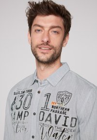 Camisa de botones a rayas grises que presenta gráficos de texto, un botón de acento naranja y un diseño de cuello pulido. Los patrones notables incluyen temas náuticos.
