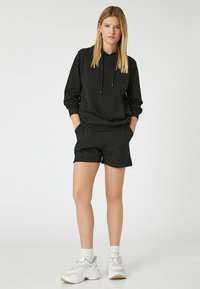 Sort hættejakke med frontlomme, parret med sorte shorts. Modellen har hvide sneakers og strømper på. Enkel, afslappet sportswear-design.