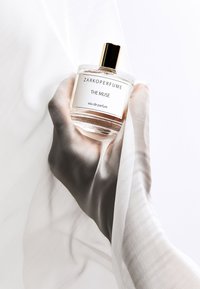 Flacon de parfum en verre transparent avec un capuchon doré. L'étiquette indique "ZARKOPERFUME THE MUSE eau de parfum." Main couverte par un tissu doux et léger.