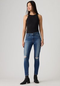 Νεαρή γυναίκα που φοράει μαύρο αμάνικο τοπ, σκισμένο στενό μπλε τζιν και μαύρα μποτάκια αστραγάλου, στέκεται μπροστά σε απλό φόντο.