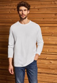 Camiseta blanca de manga larga con escote redondo y pequeño detalle de logo, combinada con unos jeans de color azul oscuro frente a un fondo de madera.
