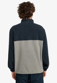 Fleece-pullover med en mörk marinblå överdel och ljusgrå underdel. Har en ståkrage och en avslappnad passform. Mjuk textur synlig.