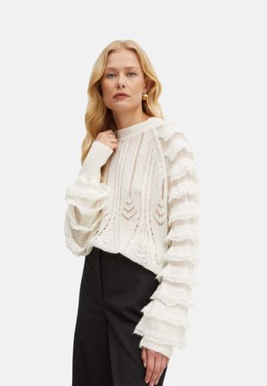 Donna bionda che indossa un maglione bianco lavorato a maglia con maniche arricciate e stratificate e pantaloni neri, in piedi davanti a uno sfondo chiaro e uniforme.