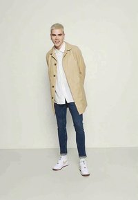 Trench coat bege claro com botões grandes, usado sobre uma camisa branca e jeans azul-escuro. Ténis brancos desportivos completam o visual.