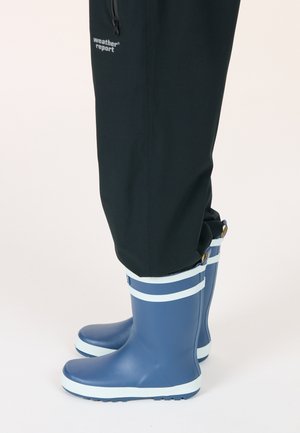 Stivali da pioggia in gomma blu con strisce bianche in cima, dotati di una suola texturizzata. Indossati con pantaloni impermeabili neri, che mettono in evidenza un design pratico.